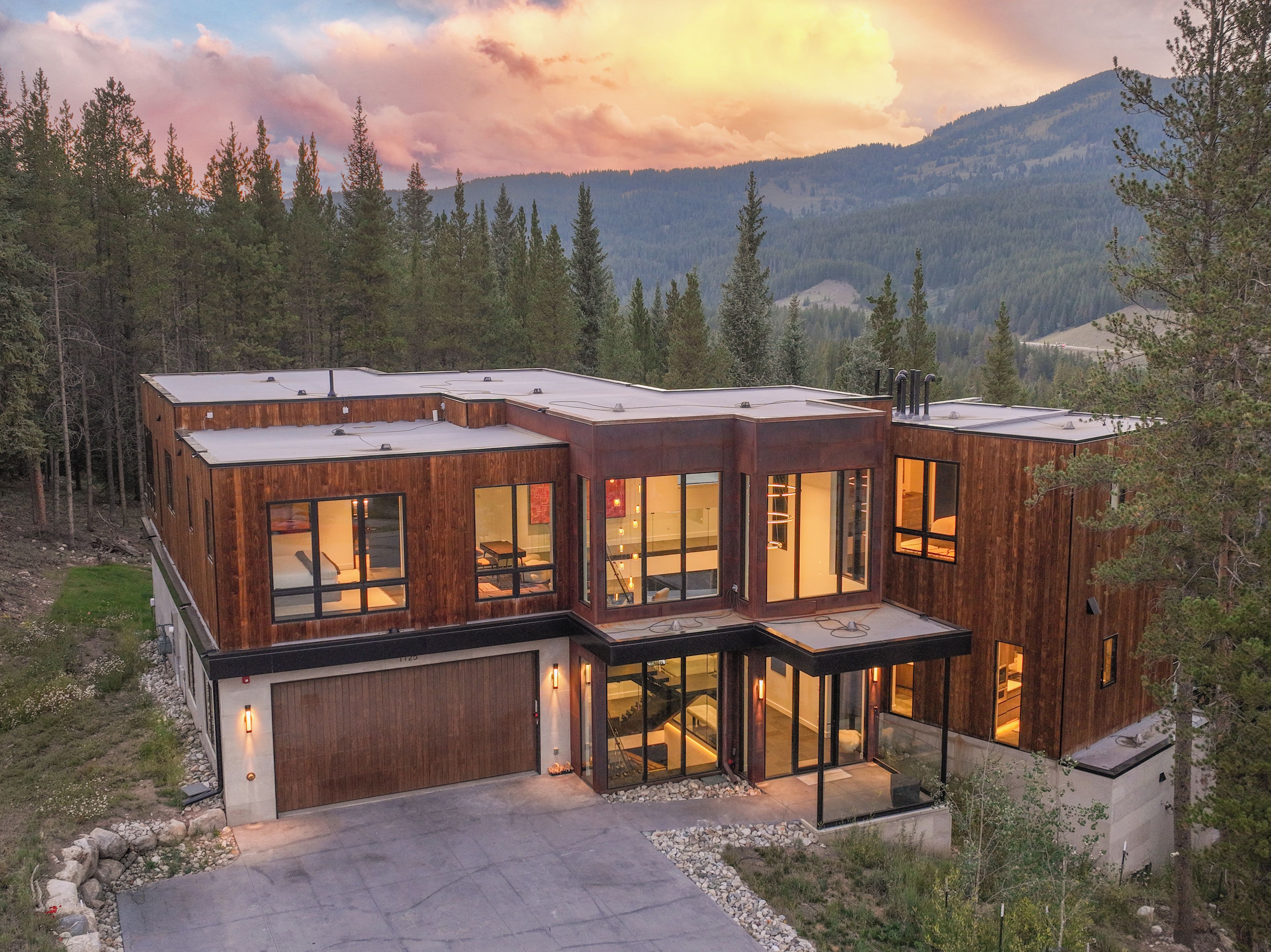 A modern wooden cabin with large windows, a garage, and a view of the surrounding forest and mountains.