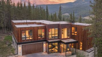 A modern wooden cabin with large windows, a garage, and a view of the surrounding forest and mountains.