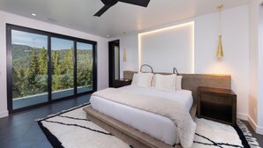 A modern bedroom with a large bed, a ceiling fan, and a view of a forest through floor-to-ceiling windows. - Steps to slopes; 7,000 sq feet of Luxury, golf simulator, hot tub! (Copper Mountain)