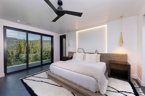 A modern bedroom with a large bed, a ceiling fan, and a view of a forest through floor-to-ceiling windows. - Steps to slopes; 7,000 sq feet of Luxury, golf simulator, hot tub! (Copper Mountain)