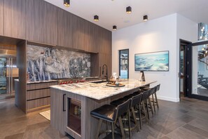 A modern kitchen with a large island, bar stools, and a view of a mountain landscape through a window. - Steps to slopes; 7,000 sq feet of Luxury, golf simulator, hot tub! (Copper Mountain)