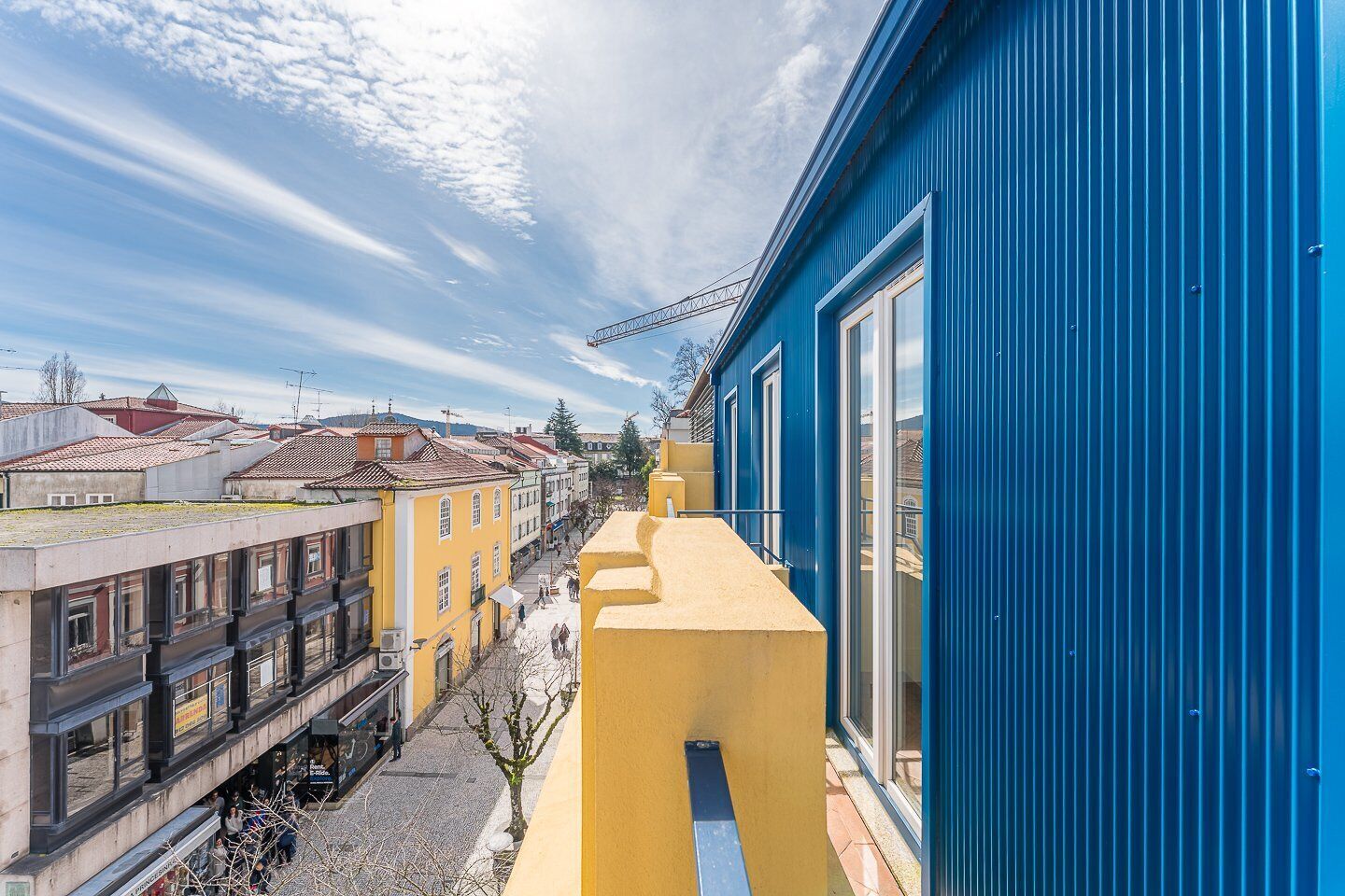 A narrow street with buildings on either side, including a blue building with large windows and a yellow building with a balcony.