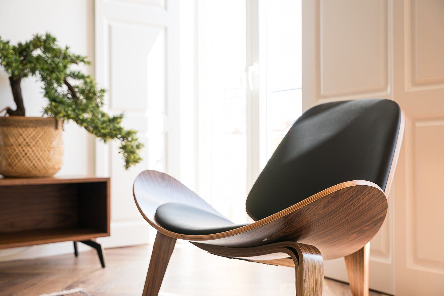 A modern chair with a dark seat and wooden frame, placed in a room with a wooden floor, a white door, and a potted plant.