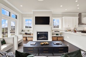 A modern kitchen with a central fireplace, white cabinetry, and a dining area with a black coffee table. - Luxury Riverfront Retreat in the Eastern Wharf | Heated Pool Access (Savannah)