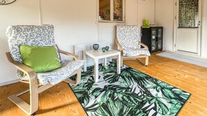 Quarto com duas cadeiras de madeira com almofadas estampadas, uma pequena mesa branca, uma planta verde e uma janela com cortinas.