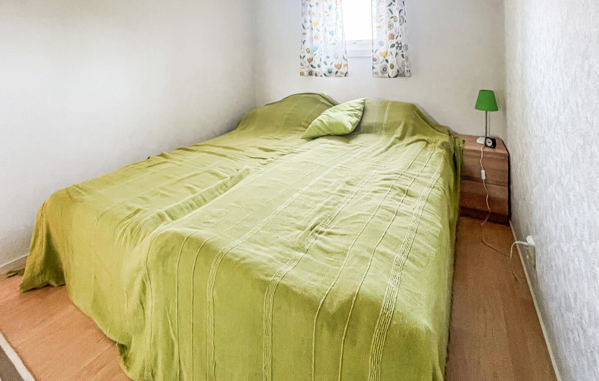 Een eenpersoonsbed met een groene sprei en kussen, een nachtkastje met een lamp en een raam met bloemen gordijnen.