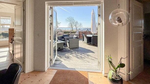 Une porte ouverte donnant sur un patio extérieur aménagé avec du mobilier et offrant une vue sur le paysage.