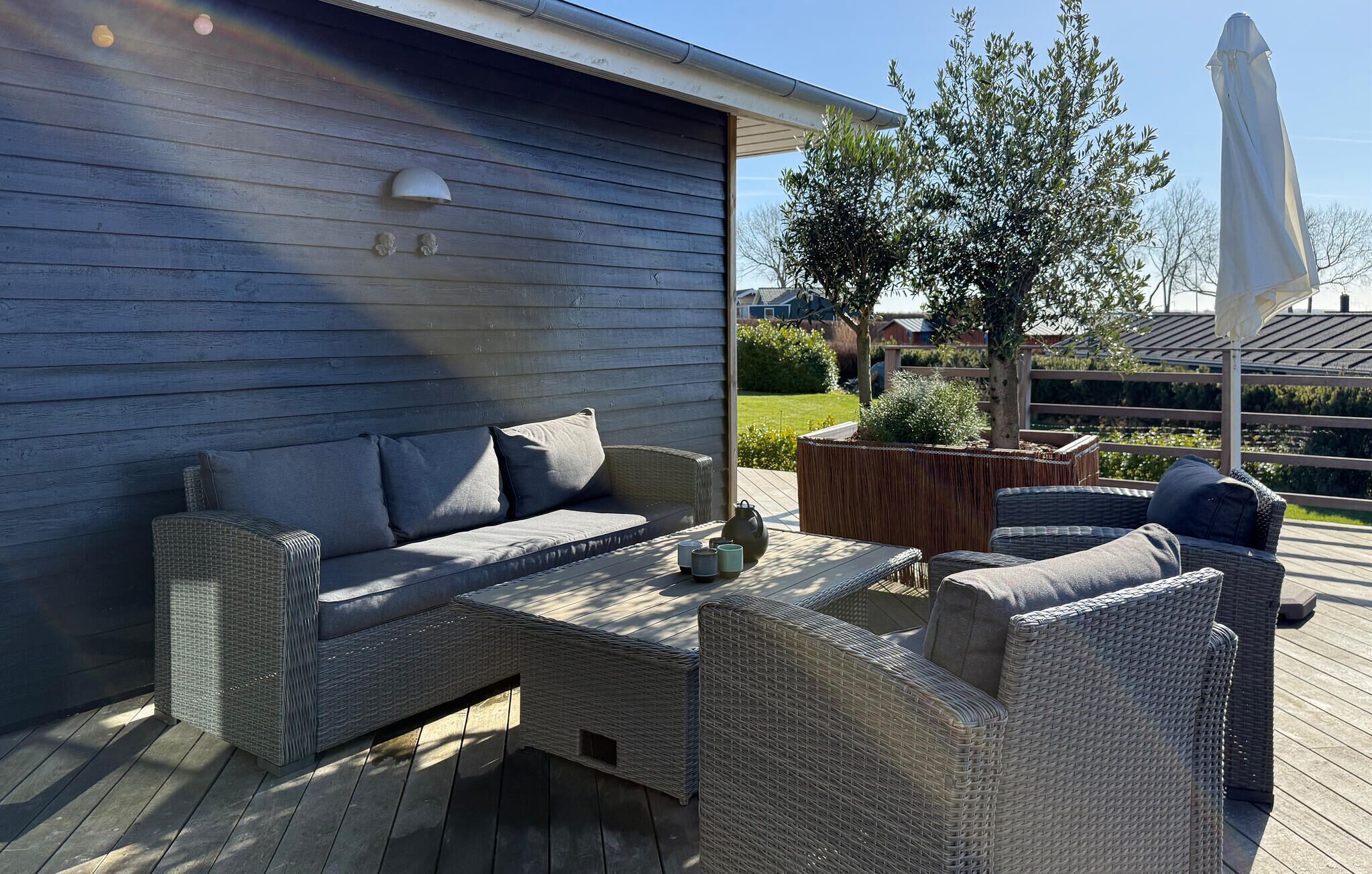 Ein überdachter Außenbereich mit Korbmöbeln, einem Tisch mit Teekanne und Blick auf einen Garten und ein Haus im Hintergrund.