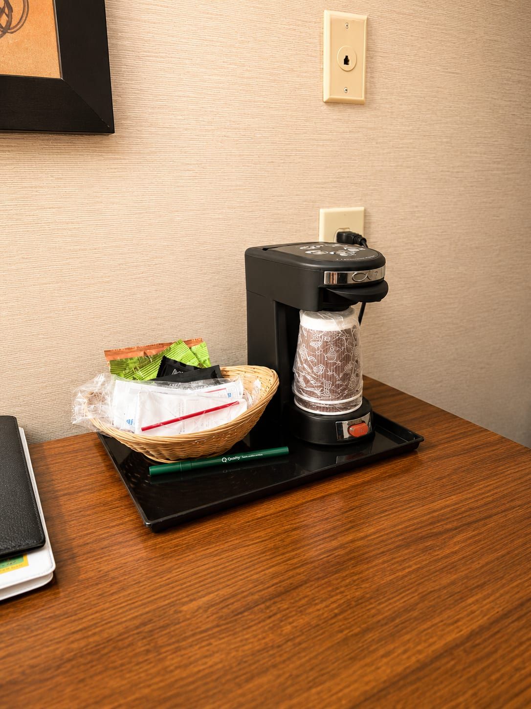 Una cafetera sobre una bandeja negra sobre una mesa de madera, una cesta con alimentos y un enchufe de pared.
