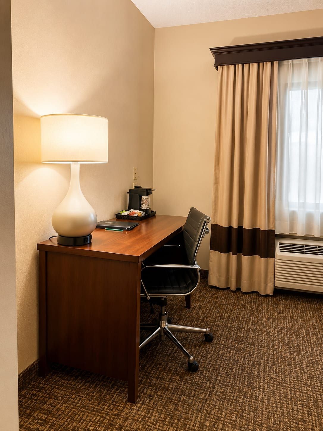 Habitación de hotel con escritorio de madera, lámpara, silla, cafetera y ventana con cortinas.