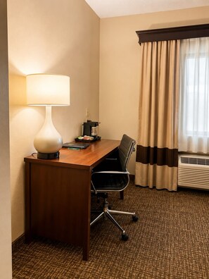 Habitación de hotel con escritorio de madera, lámpara, silla, cafetera y ventana con cortinas.