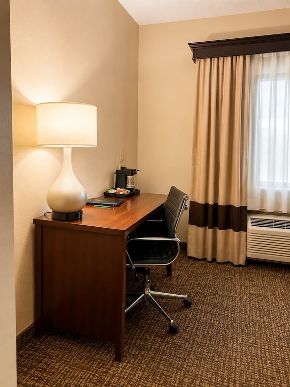 Une chambre d’hôtel avec un bureau en bois, une lampe, une chaise, une machine à café et une fenêtre avec des rideaux.