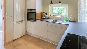 Eine kompakte Küche mit Kühlschrank, Backofen, Kaffeemaschine und einem Fenster mit Blick ins Grüne.