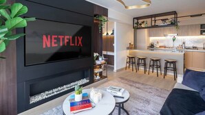 A modern living room with a large flat-screen TV, a fireplace, a round coffee table with books and candles, and a kitchen area with bar stools. - Luxury Boutique Apt | Miraflores | Ocean View (Miraflores)
