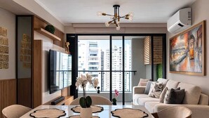 Un salon moderne doté d’un téléviseur à écran plat, d’une table à manger avec des sets de table, d’un canapé et d’une grande fenêtre offrant une vue sur la ville.