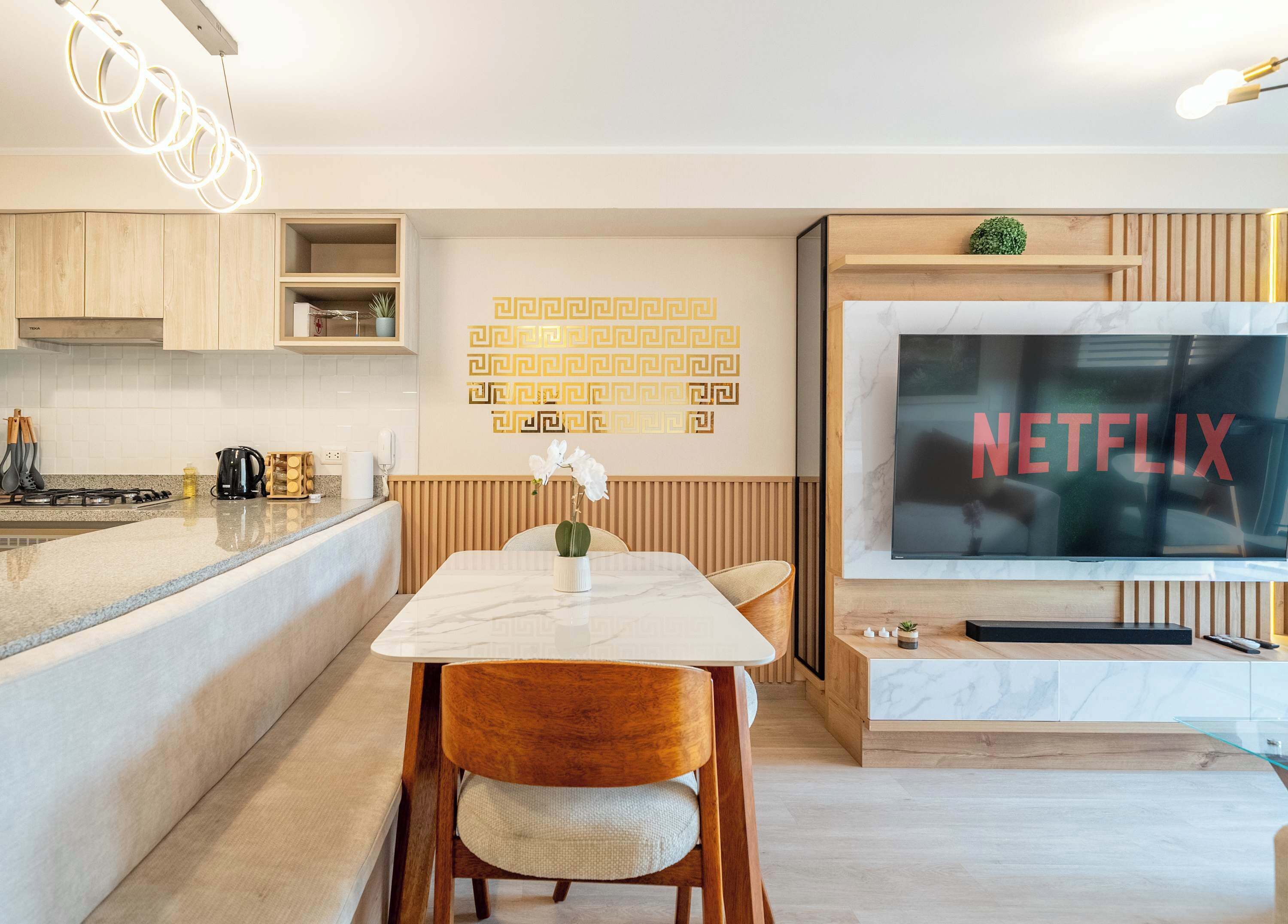 Una cocina moderna con encimera de mármol, armarios de madera, horno integrado y comedor con mesa y sillas blancas.