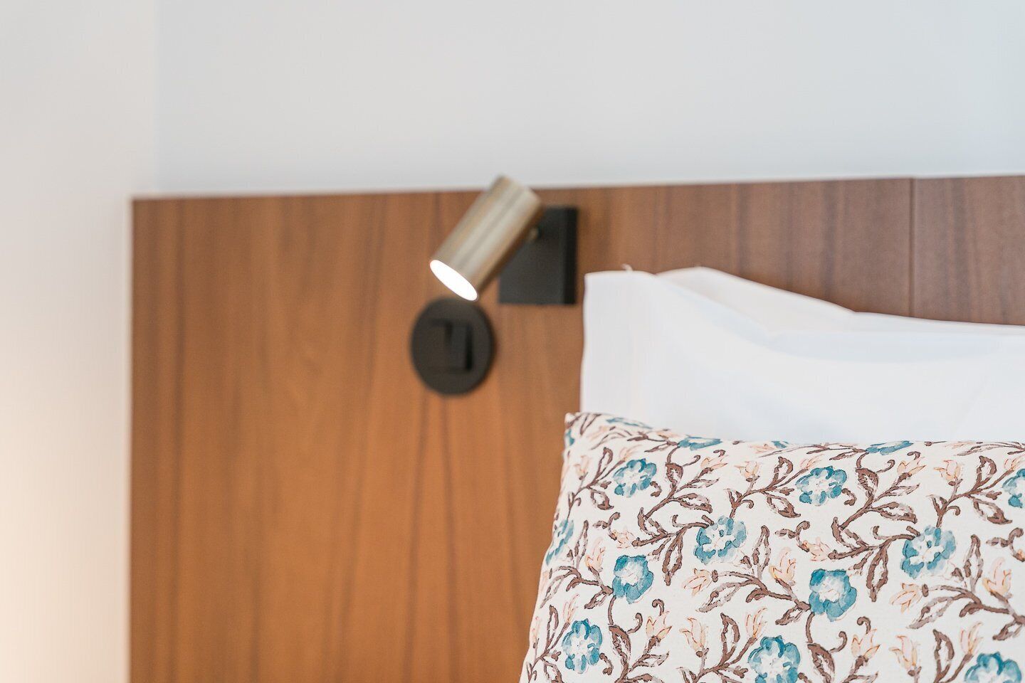 Una cama con un cojín estampado y una lámpara fijada en la pared.