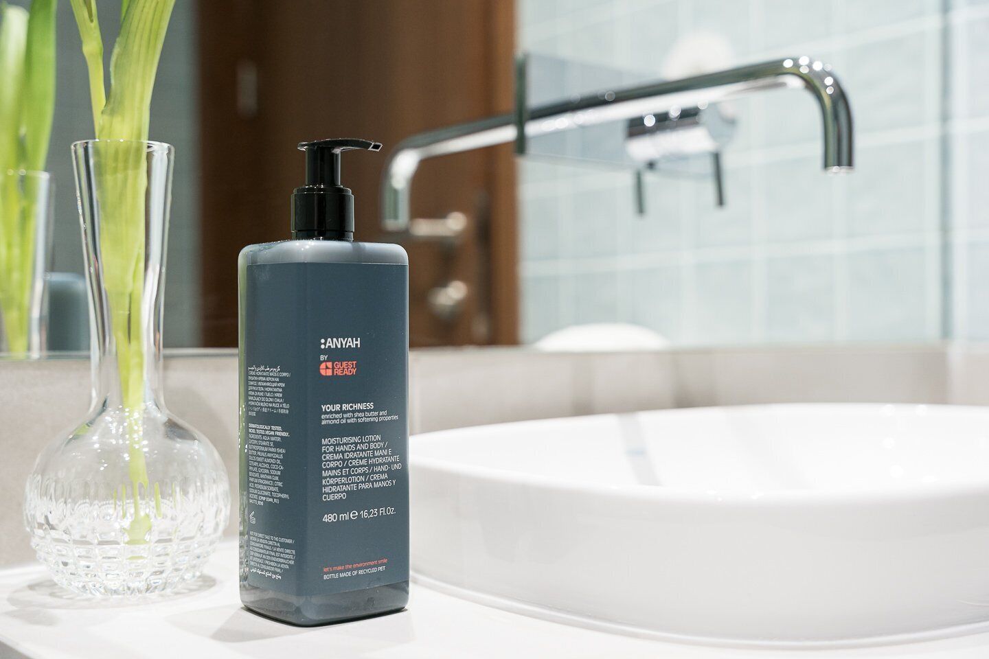A bathroom with a modern sink, a clear glass vase with green leaves, and a bottle of AnyaH by Guest Ready moisturizing lotion.