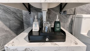 Un baño con encimera de mármol, una bandeja negra que contiene una botella de gel de ducha, una botella de champú y un recipiente de vidrio.