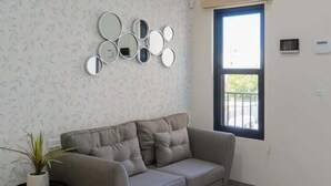 A living room with a grey sofa, a small side table with a plant, a window with blinds, and a wall-mounted mirror arrangement. - Pool, BBQ & a Short Walk to the Malecon (La Paz)