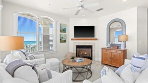A living room with a fireplace, white furniture, a ceiling fan, and a large window with a view. - Gulf Front w/ Amazing Panoramic Gulf Views! Seacrest Beach | Private Beach Access (Seacrest Beach)