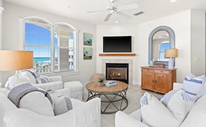 A living room with a fireplace, white furniture, a ceiling fan, and a large window with a view. - Gulf Front w/ Amazing Panoramic Gulf Views! Seacrest Beach | Private Beach Access (Seacrest Beach)