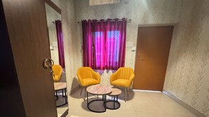 A room with two yellow chairs, a small round table, a mirror, a door, and patterned wallpaper. - Devashish Homestay (Varanasi)
