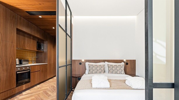 A modern hotel room with a wooden floor, a bed with white linens, a built-in kitchen with a microwave, and a large window. - GuestReady - Santa Barbara Garden House 12 (Braga)