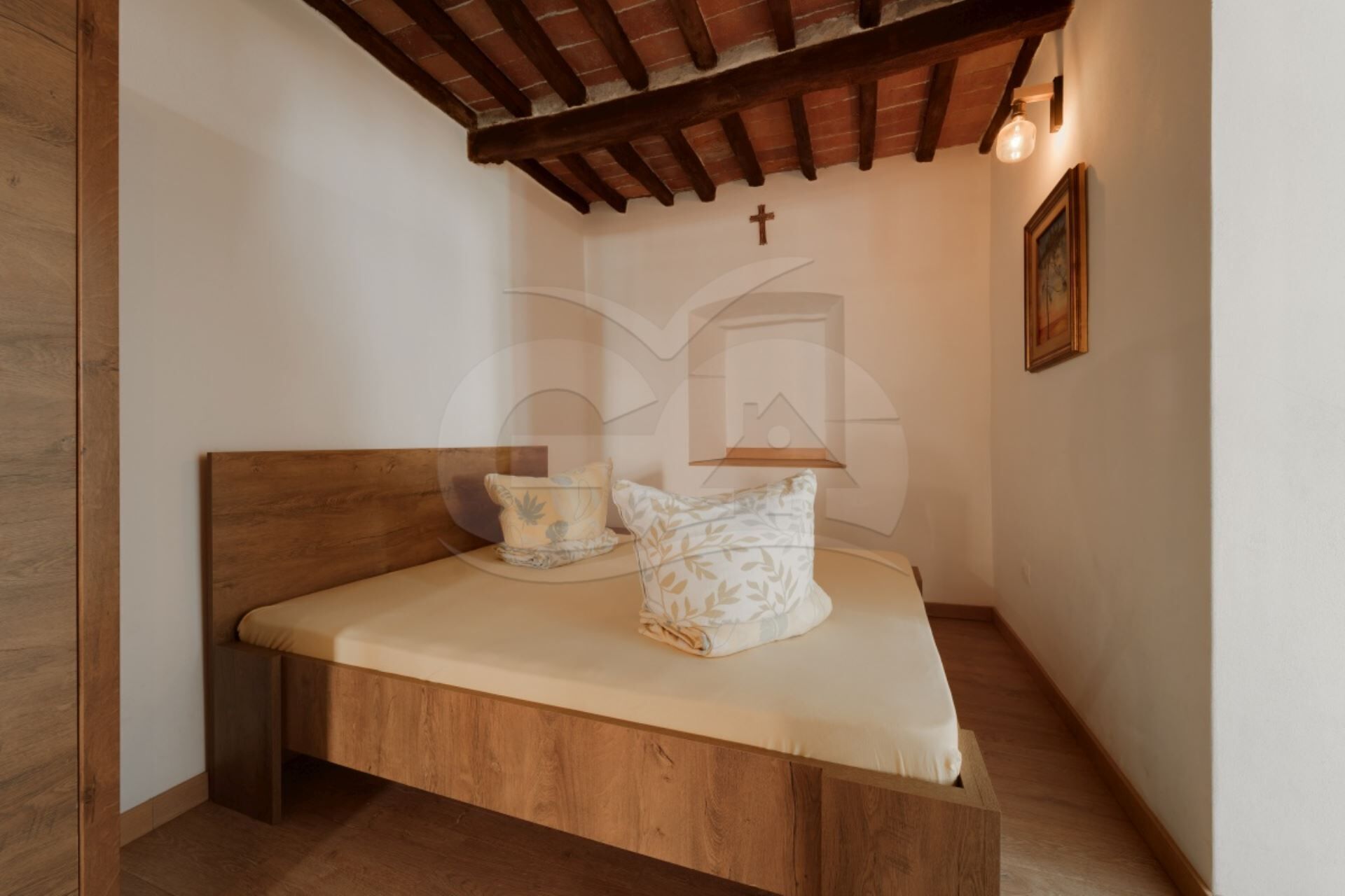 A small, cozy room with a wooden bed, a cross on the wall, and a framed picture.