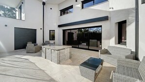 A modern outdoor patio with wicker furniture, a glass-top table, and a garage door. - Luxury Villa in Grimaud (GRIMAUD)