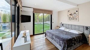 A bedroom with a bed, a TV, a desk, and a view of a garden. - Luxury Villa in Grimaud (GRIMAUD)