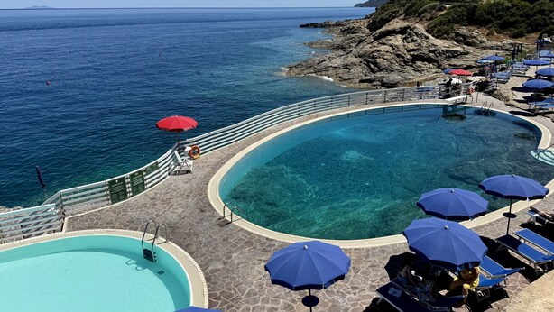 Uma área de piscina com água azul cristalina, espreguiçadeiras e guarda-sóis.