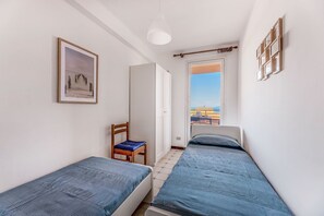 Ett litet, ljust rum med två enkelsängar, en stol, en inramad tavla och utsikt över havet genom en dörr.