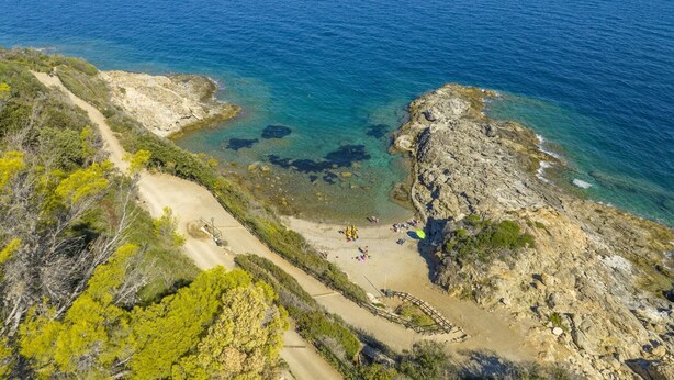 Eine Küstenlandschaft mit klarem, blauem Meer, felsigem Gelände und einem Feldweg, der zu einem Strand führt.
