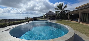 A swimming pool with a curved edge, a thatched-roof structure, palm trees, and a view of the ocean. - Villa with Pool & Jacuzzi, 180° panoramic view of the sea and Moorea (Punaauia)