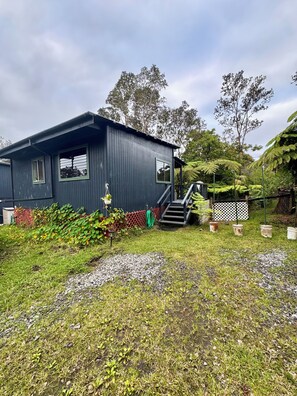 A single-story building with a dark exterior, a small porch, and a staircase leading to the entrance. - Rainforest Studio in Volcano- 3miles from National Park (Volcano)