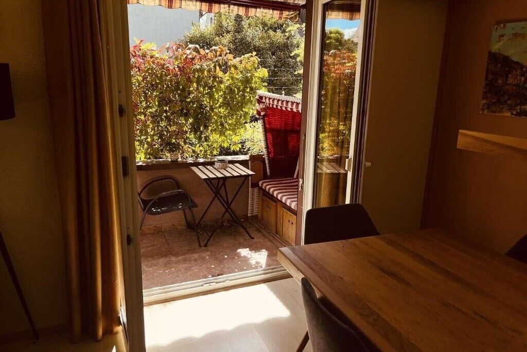 Une pièce comprenant une table en bois, une chaise, une petite table avec une chaise, et donnant sur un jardin avec des arbres et une structure rouge.