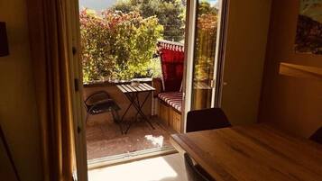 Une pièce comprenant une table en bois, une chaise, une petite table avec une chaise, et donnant sur un jardin avec des arbres et une structure rouge.