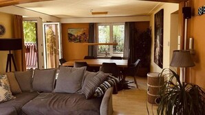 A cozy living room with a sofa, dining table, and chairs. There is a window with curtains, a lamp, and a clock on the wall. - Centurio 5 :Double room with balcony (Augst)