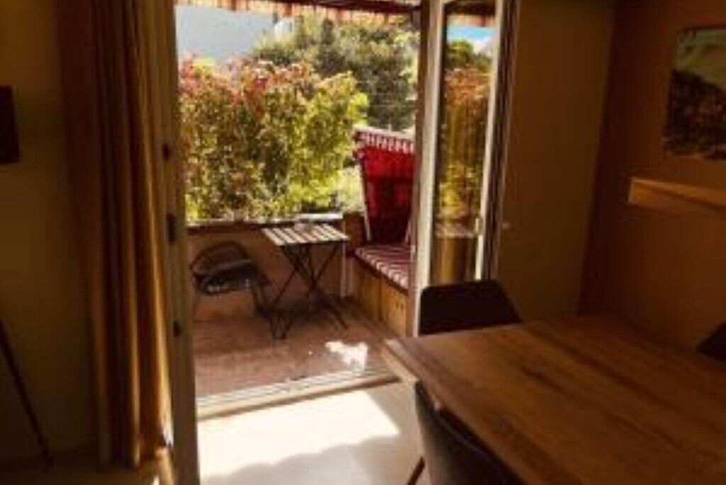 A room with a wooden table, a window with a view of greenery, and a red chair outside.
