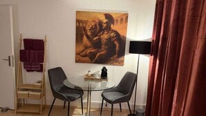 A room with a painting of a historical figure, a small round table with chairs, a lamp, and a wooden shelf with baskets. - Centurio 5 :Double room with balcony (Augst)