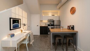Et kompakt køkken med moderne hårde hvidevarer, et spiseområde med barstole og et lille arbejdsområde med skrivebord og stol.