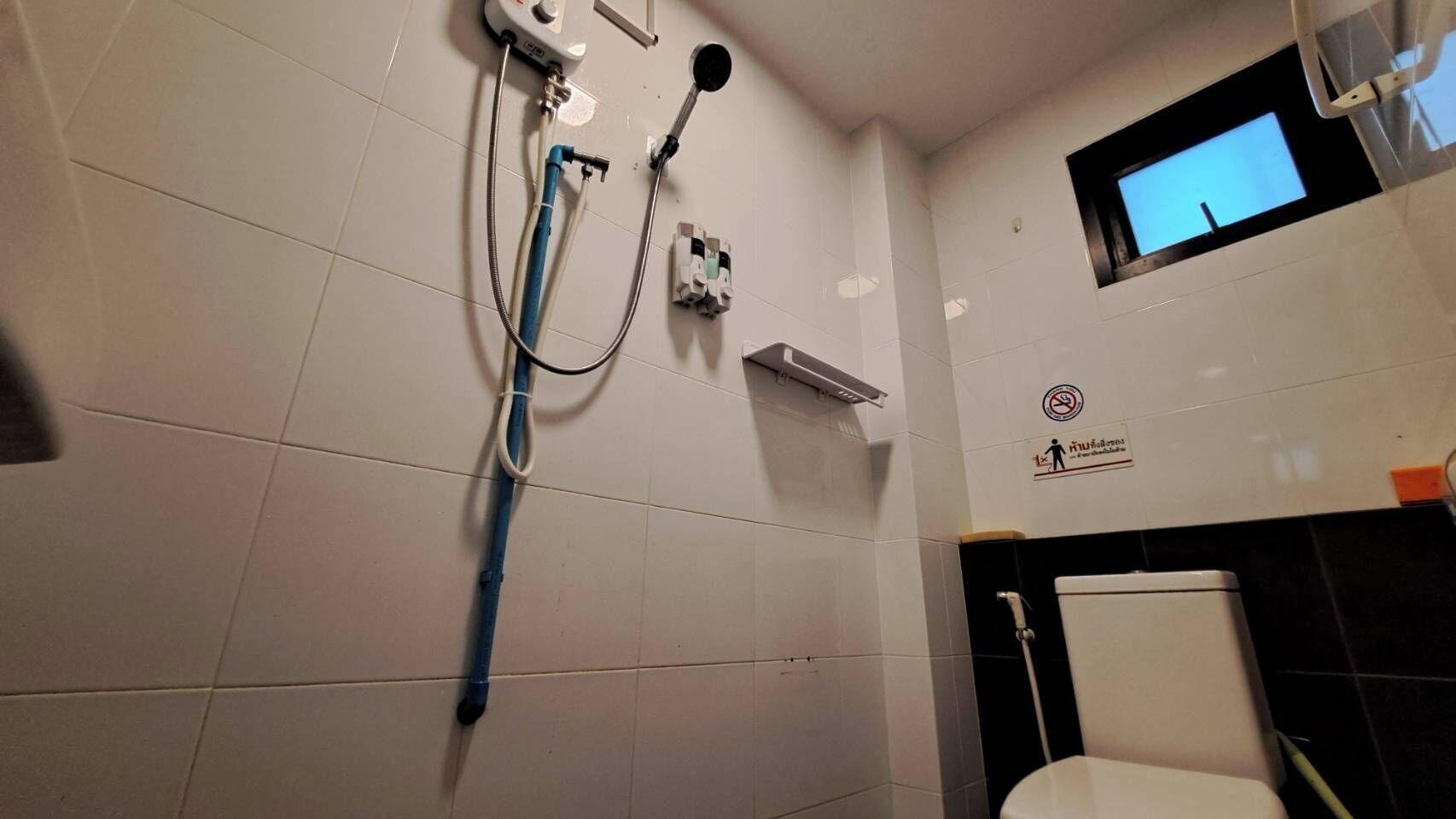 Ein Badezimmer mit weißen Fliesen, einer Dusche inklusive Handbrause, einer Toilette und einem kleinen Fenster.