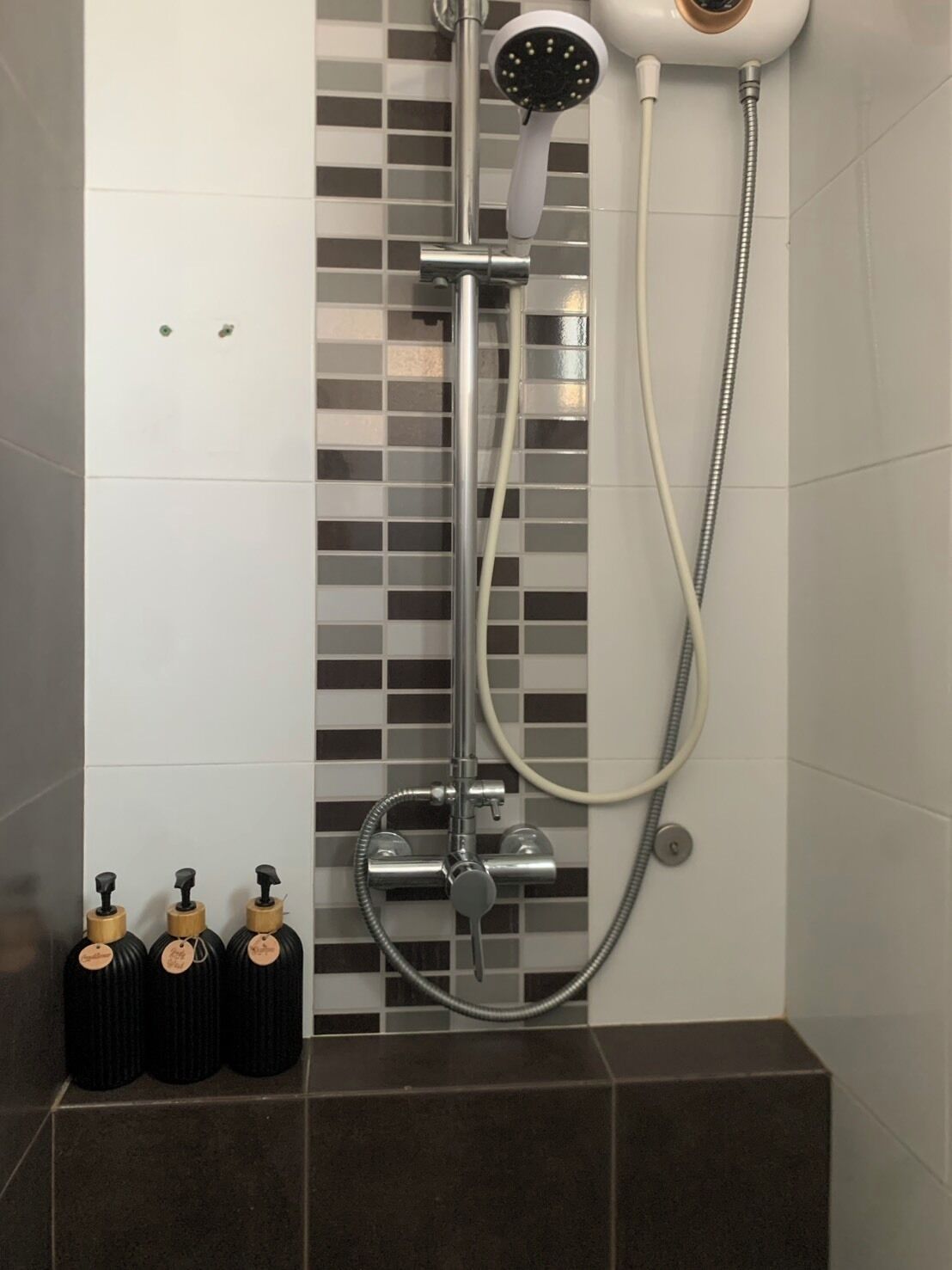 A shower with a handheld showerhead, a wall-mounted faucet, and a handheld hose. There are three black bottles with wooden caps on the floor.