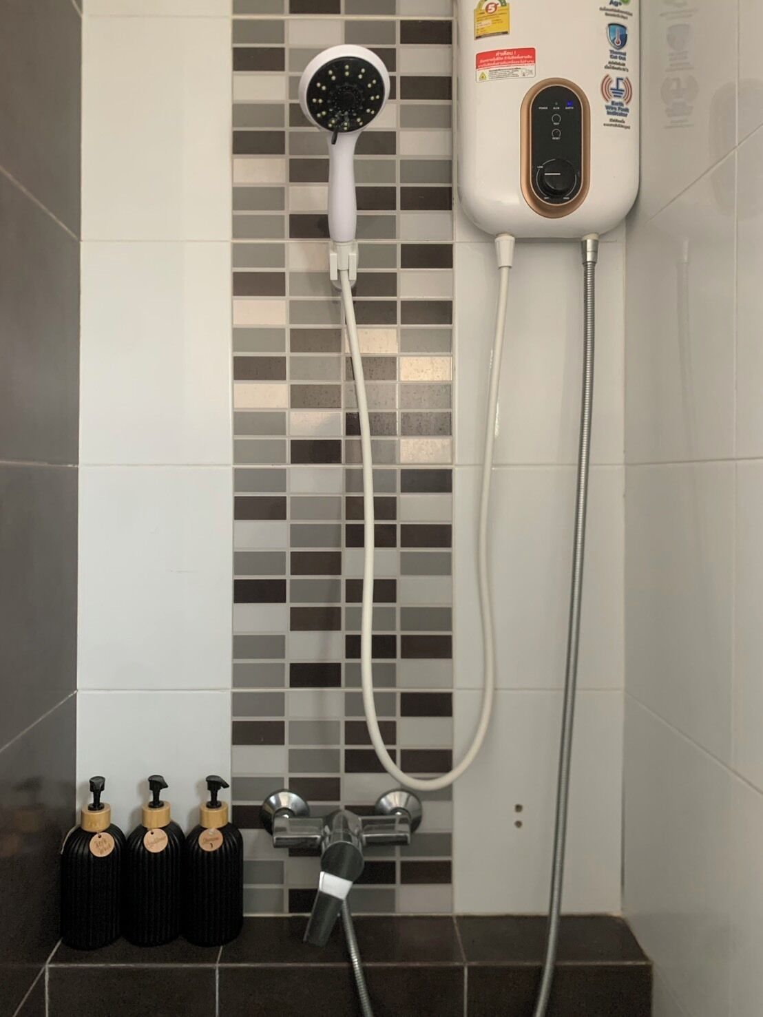 A shower with a handheld showerhead, a wall-mounted control panel, and three black bottles with pumps on a tiled wall.