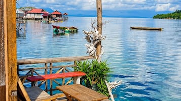 A wooden deck with a bench and table overlooking a calm body of water, with a wooden post and plants in the foreground.