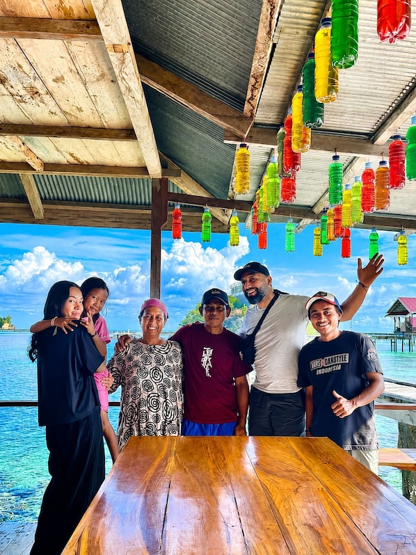A group of people standing at a wooden table with a view of the ocean and a clear sky.