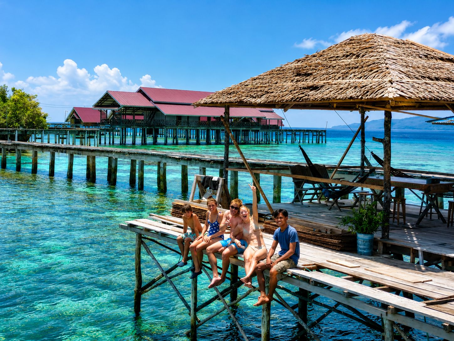 A wooden pier extending into clear blue water with a thatched-roof structure and a building with a red roof in the background.
