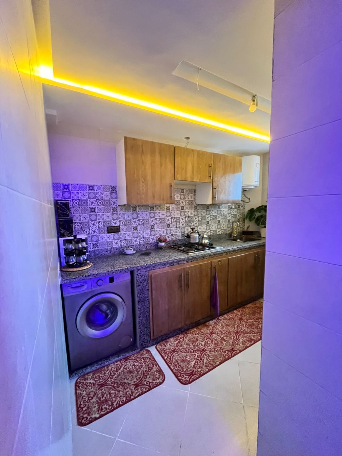 Une cuisine compacte équipée d’un lave-linge, de meubles en bois, de carreaux décoratifs et d’un luminaire moderne.