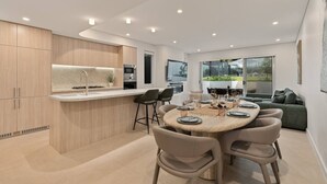 A modern kitchen with a central island, dining area with chairs, and a large window. - Bayside Luxury Retreat with Pool Near to Cronulla (Port Hacking)